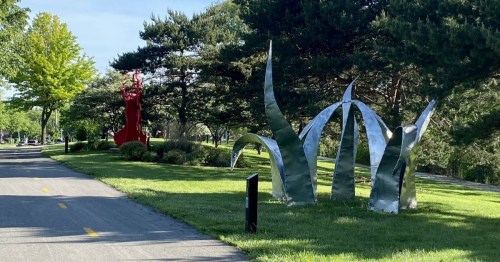 A scenic pathway lined with large metallic sculptures resembling grass and a red abstract figure, surrounded by trees.