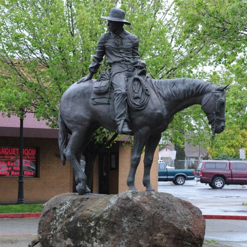 Bronze statue of a cowboy on horseback, sitting on a rock, surrounded by green trees and a building in the background.