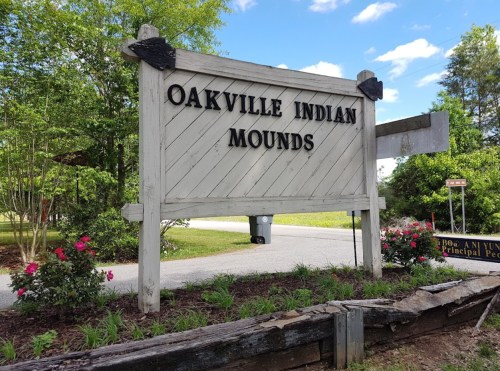 Sign for Oakville Indian Mounds, surrounded by greenery and flowers, with a road visible in the background.