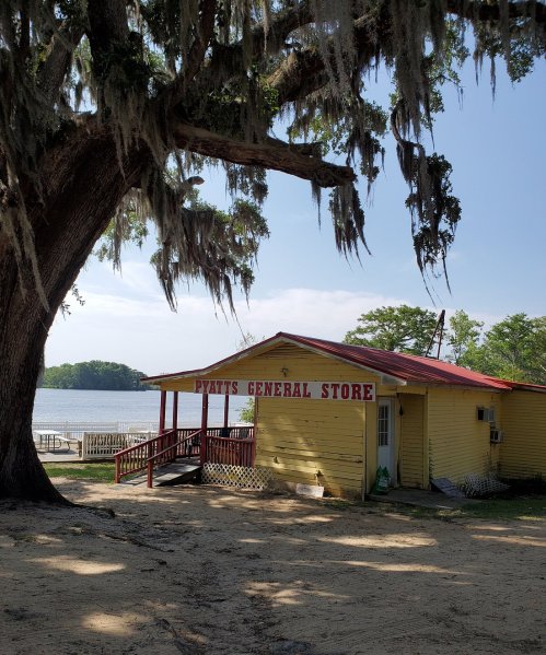 A yellow general store with a red roof, surrounded by trees and near a lake under a clear blue sky.
