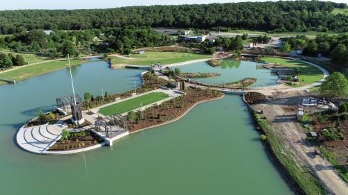 Aerial view of a landscaped park with pathways, water features, and greenery, surrounded by trees and open space.