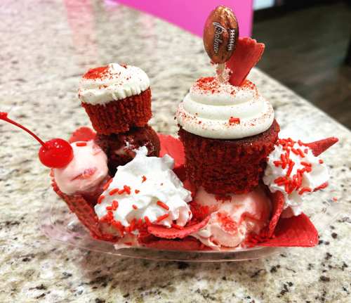 A colorful dessert featuring red velvet cupcakes, ice cream, whipped cream, and festive sprinkles in a clear cup.