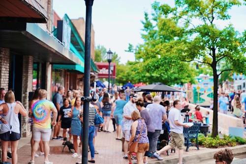 A busy street scene with a crowd of people walking, shopping, and socializing outside various shops and cafes.