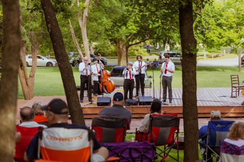 A band performs on stage in a park while an audience sits in folding chairs, surrounded by trees.