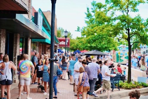 A busy street scene with a crowd of people walking, shopping, and socializing outside various shops and cafes.