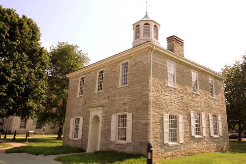 Historic stone building with a cupola, surrounded by greenery and trees under a clear blue sky.