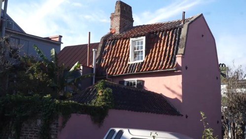 A pink house with a tiled roof and chimney, surrounded by greenery and a parked car in the foreground.