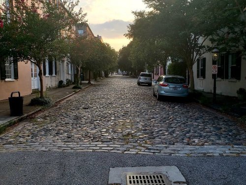 A quiet, cobblestone street lined with trees and parked cars, under a soft, golden sunset.
