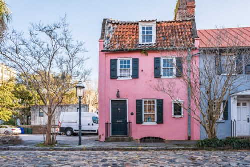 A charming pink house with a tiled roof, black shutters, and a lantern, surrounded by trees and cobblestone streets.