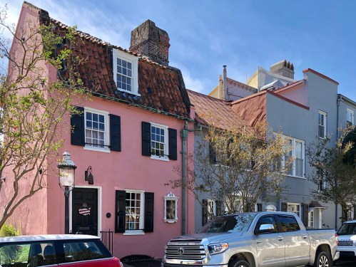A pink house with black shutters next to a gray house, with a truck parked in front and trees nearby.