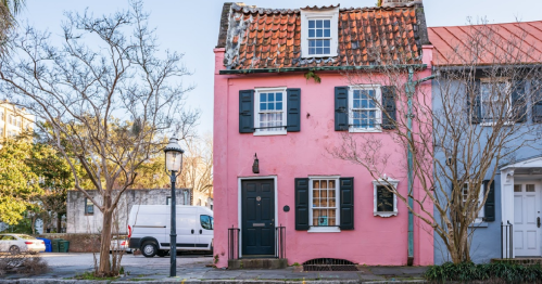 A charming pink house with a tiled roof and black shutters, surrounded by trees and a streetlamp.