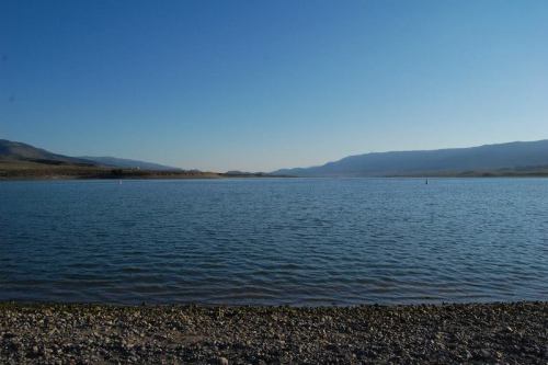 A serene lake surrounded by mountains under a clear blue sky, with a rocky shoreline in the foreground.