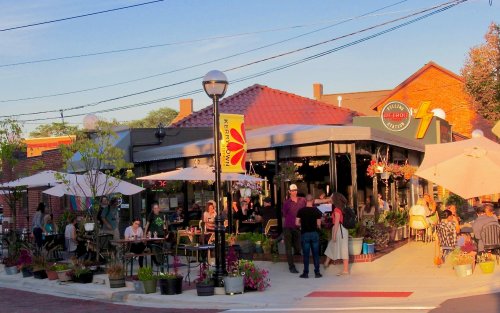 A lively outdoor café scene with people dining, surrounded by plants and umbrellas, under a clear blue sky.