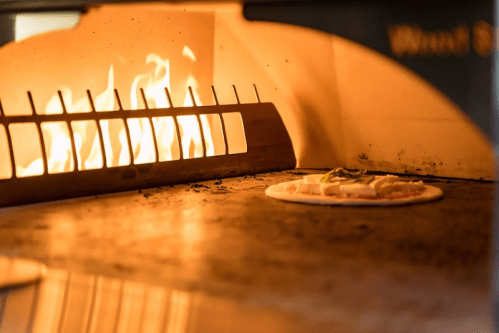 A pizza baking in a wood-fired oven with flames visible in the background.