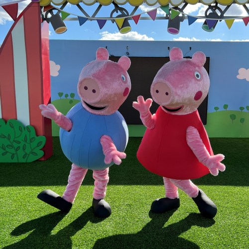 Two cartoon pig characters in colorful outfits stand cheerfully on a grassy stage with a festive backdrop.