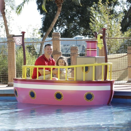 A man and a child smile while sitting on a colorful boat in a pool surrounded by palm trees.