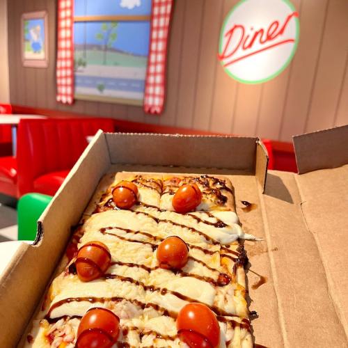 A pizza in a takeout box topped with cherry tomatoes, set in a retro diner with red booths and checkered curtains.