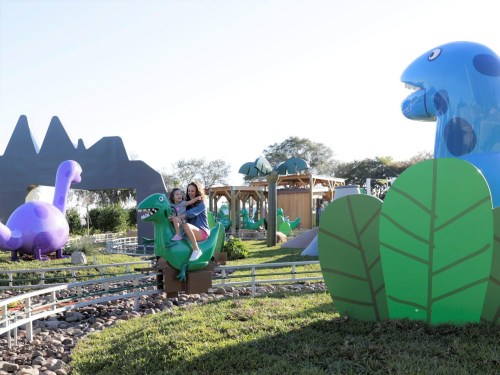 Two children play on a dinosaur sculpture in a colorful outdoor park with large, whimsical dinosaur figures in the background.