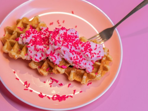 A waffle topped with pink whipped cream and pink sprinkles, served on a pastel pink plate with a fork.