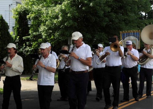 A marching band in white shirts and hats plays instruments while parading down a tree-lined street.