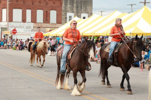 Three riders on horseback in orange shirts parade down a street lined with spectators and a large yellow tent.