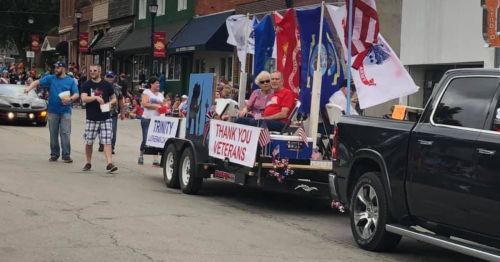 A parade float with veterans, flags, and a "Thank You Veterans" sign, surrounded by a cheering crowd.