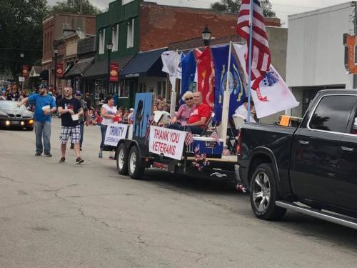 A parade float with veterans, flags, and a "Thank You Veterans" sign, moving through a small town street.