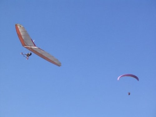 Two gliders soaring in a clear blue sky, one with a rigid wing and the other with a parachute-like canopy.