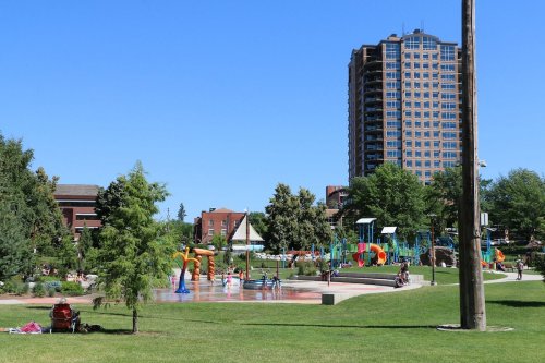 A sunny park with a splash pad, playground equipment, and a tall building in the background. Trees and people are visible.