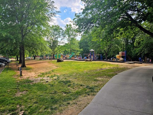 A playground surrounded by trees, with colorful equipment and children playing, viewed from a nearby path.