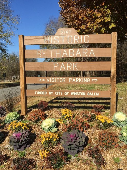 Wooden sign for Historic Bethabara Park, indicating visitor parking, surrounded by colorful flowers. Funded by the city.