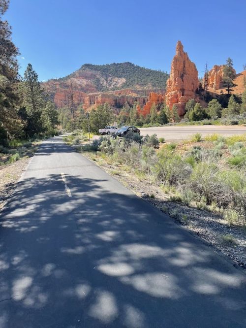 A paved path leads through greenery, with red rock formations and a blue sky in the background.