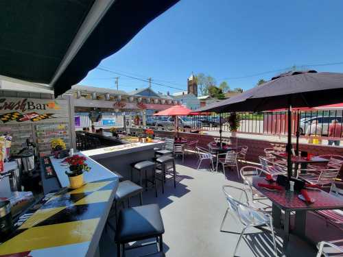 Outdoor dining area with colorful umbrellas, tables, and a bar under a clear blue sky. Flowers add a vibrant touch.