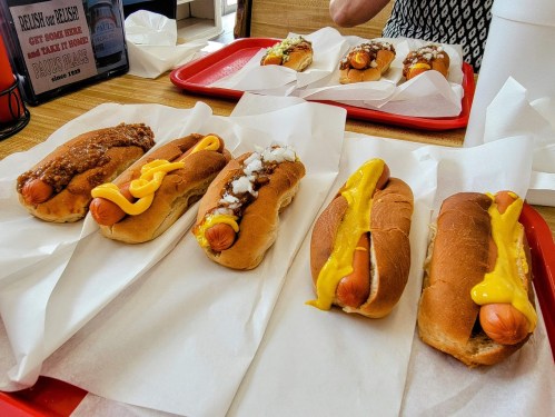 A tray of six hot dogs with various toppings, including chili, onions, and mustard, on a table.