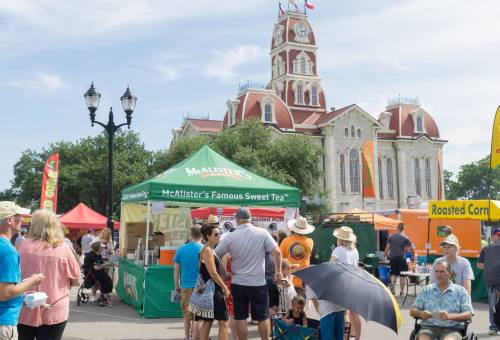 A busy outdoor festival scene with food stalls and a historic building in the background under a clear blue sky.