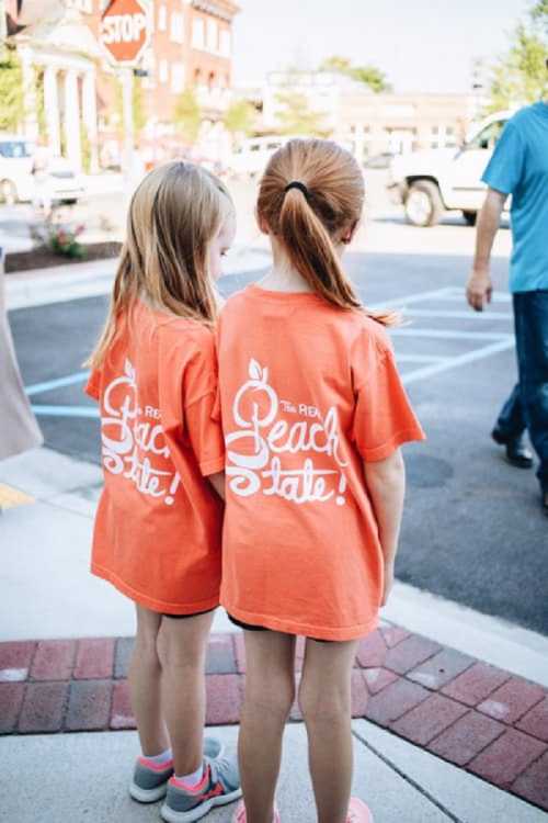 Two girls in matching orange t-shirts stand back-to-back, facing away, in a sunny outdoor setting.