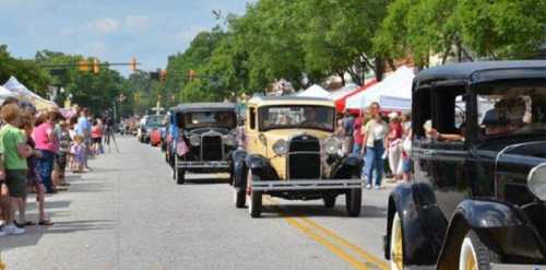 A parade of vintage cars drives down a street lined with spectators and colorful tents under a blue sky.