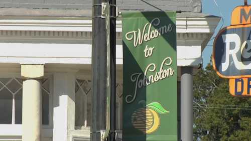 A green sign reading "Welcome to Johnston" with an orange graphic, set against a building backdrop.