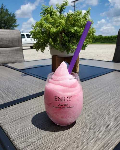 A pink frozen drink in a glass with a purple straw, placed on a table outdoors with a potted plant in the background.