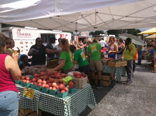 A bustling farmers market with people shopping for fresh peaches and other produce under a white tent.