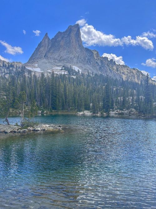 A serene lake surrounded by tall trees and a majestic mountain under a blue sky with fluffy clouds.