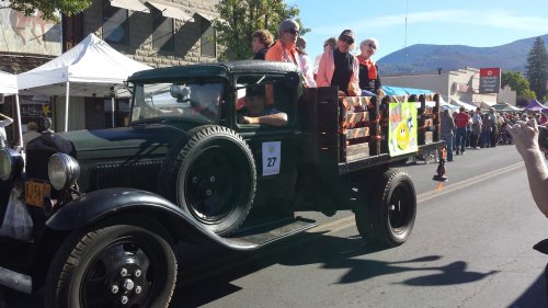 A vintage black truck in a parade, carrying people and colorful decorations, with a festive atmosphere and blue sky.