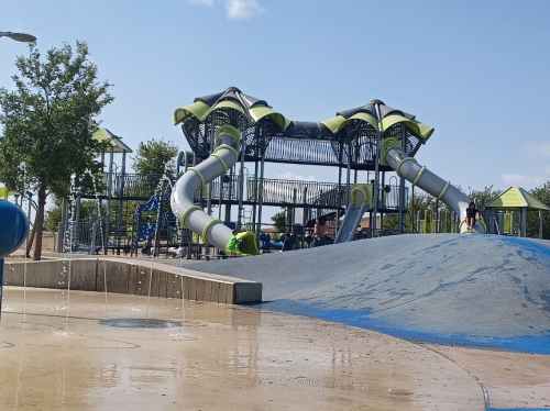 A colorful playground with slides and climbing structures, surrounded by trees and a splash pad in the foreground.