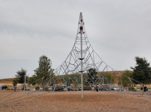 A large climbing structure made of metal ropes in a playground, with children playing and trees in the background.