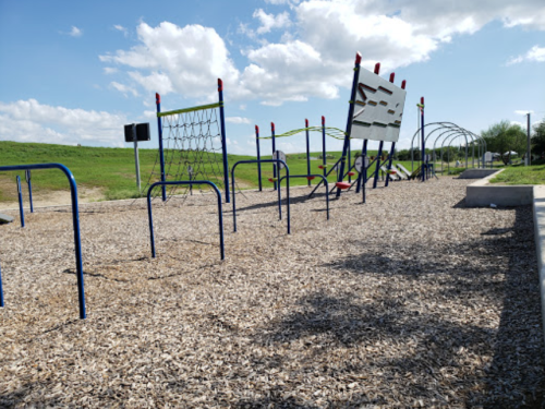 A playground with climbing structures and equipment on a sunny day, surrounded by grassy hills and blue skies.