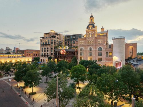 Historic buildings and modern architecture surround a tree-filled plaza at dusk, with a food hall and hotel visible.