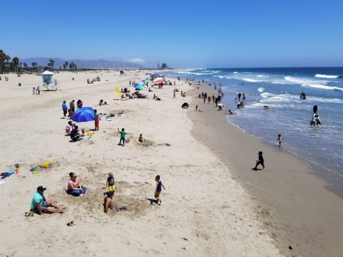 A sunny beach scene with people relaxing, playing, and walking along the shore, with umbrellas and waves in the background.