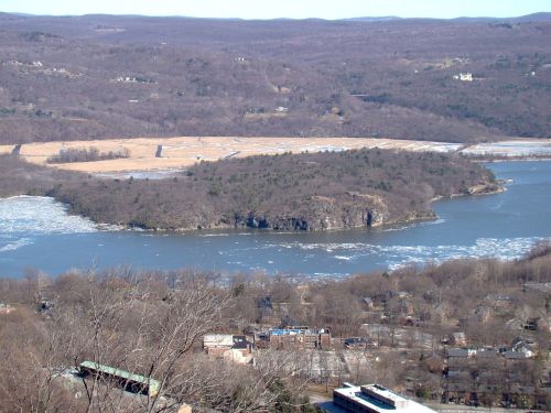 A scenic view of a river winding through a landscape with ice, trees, and distant hills under a clear sky.