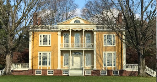 A large yellow historic house with white trim, featuring a balcony and surrounded by bare trees against a blue sky.