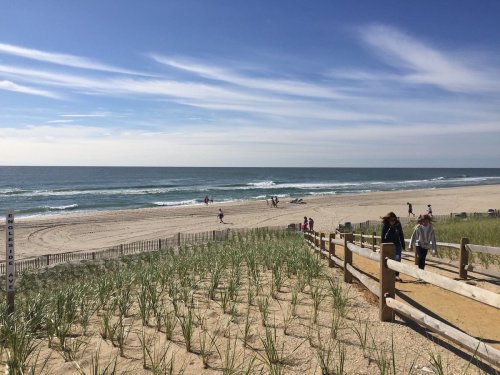 A sunny beach scene with people walking along the shore and gentle waves under a blue sky with wispy clouds.
